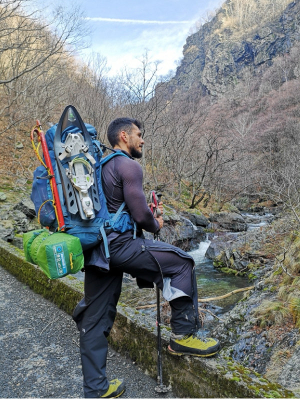 Даниел Иванов по време на планински преход край река, екипиран с голяма туристическа раница с прикрепени към нея снегоходки, пикел и палатка, облечен в професионално облекло за планинарство.