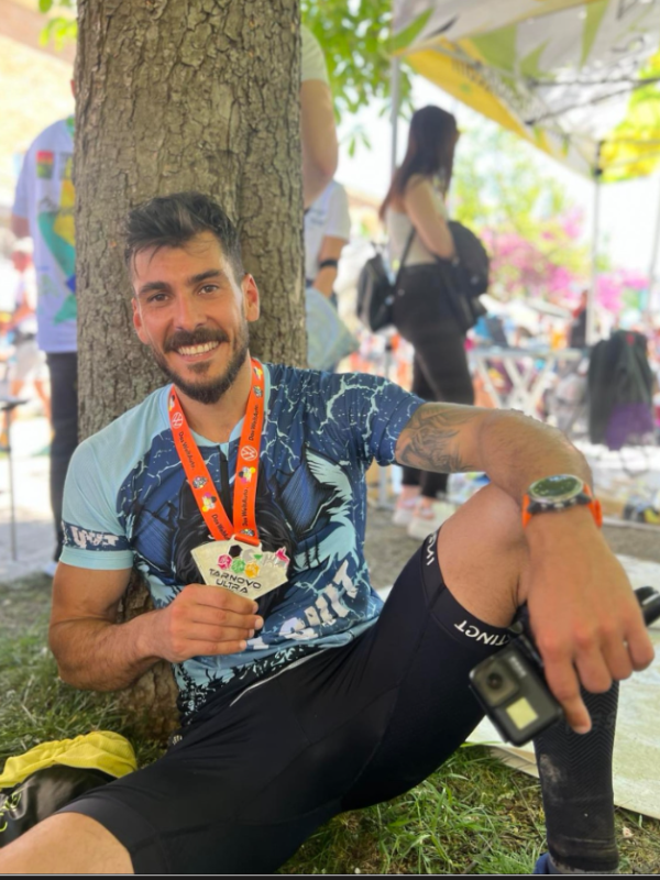 Усмихнатият Даниел Иванов след финала на състезанието Tarnovo Ultra, показващ своя медал. Той е облечен в спортна тениска за бягане и клин, седнал под дърво в зоната за почивка.