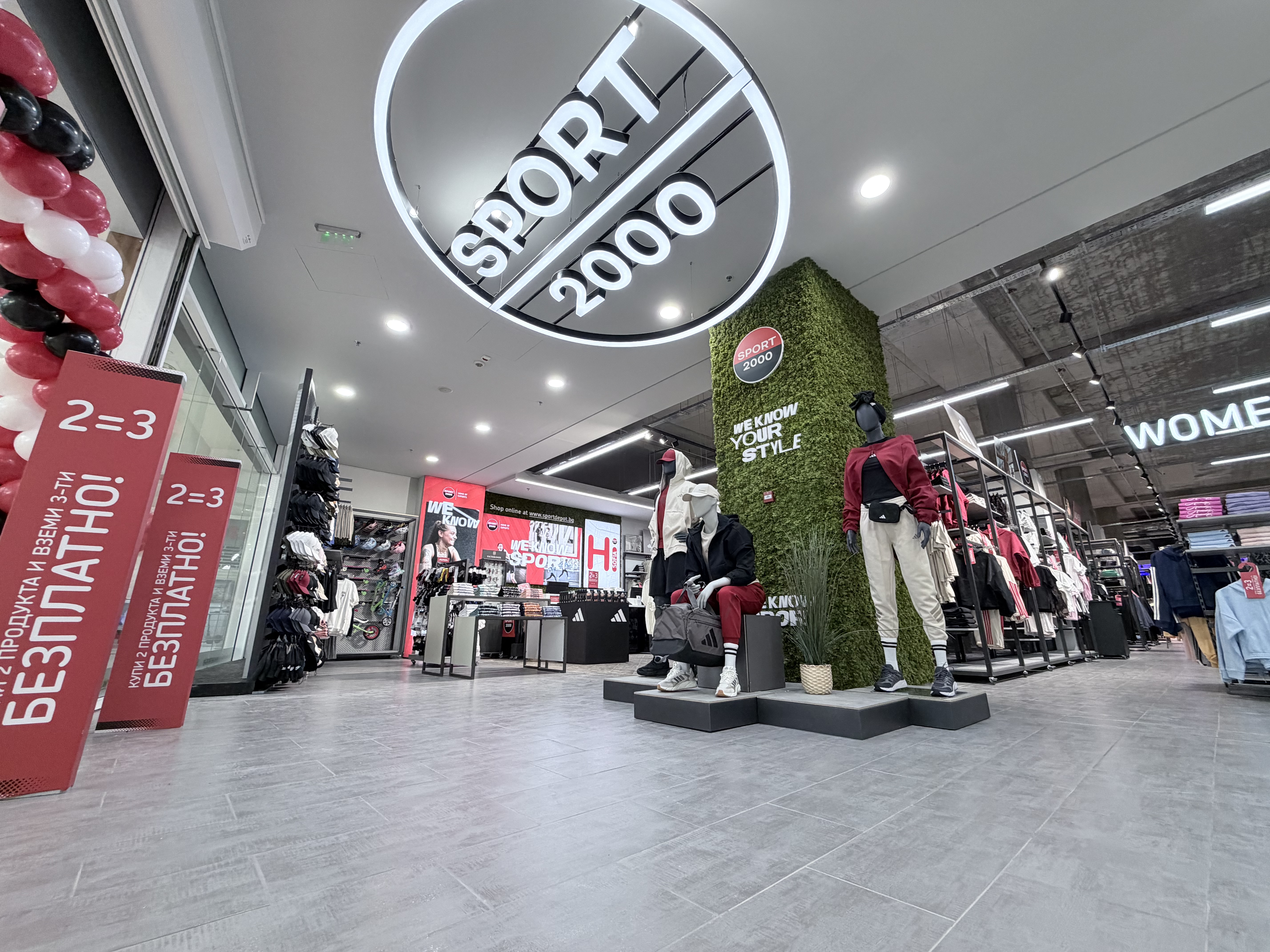 Изглед отвътре на SPORT DEPOT със зелена стена тип &bdquo;жива картина&ldquo;, стилни манекени в спортно-ежедневен стил и модерно осветление.