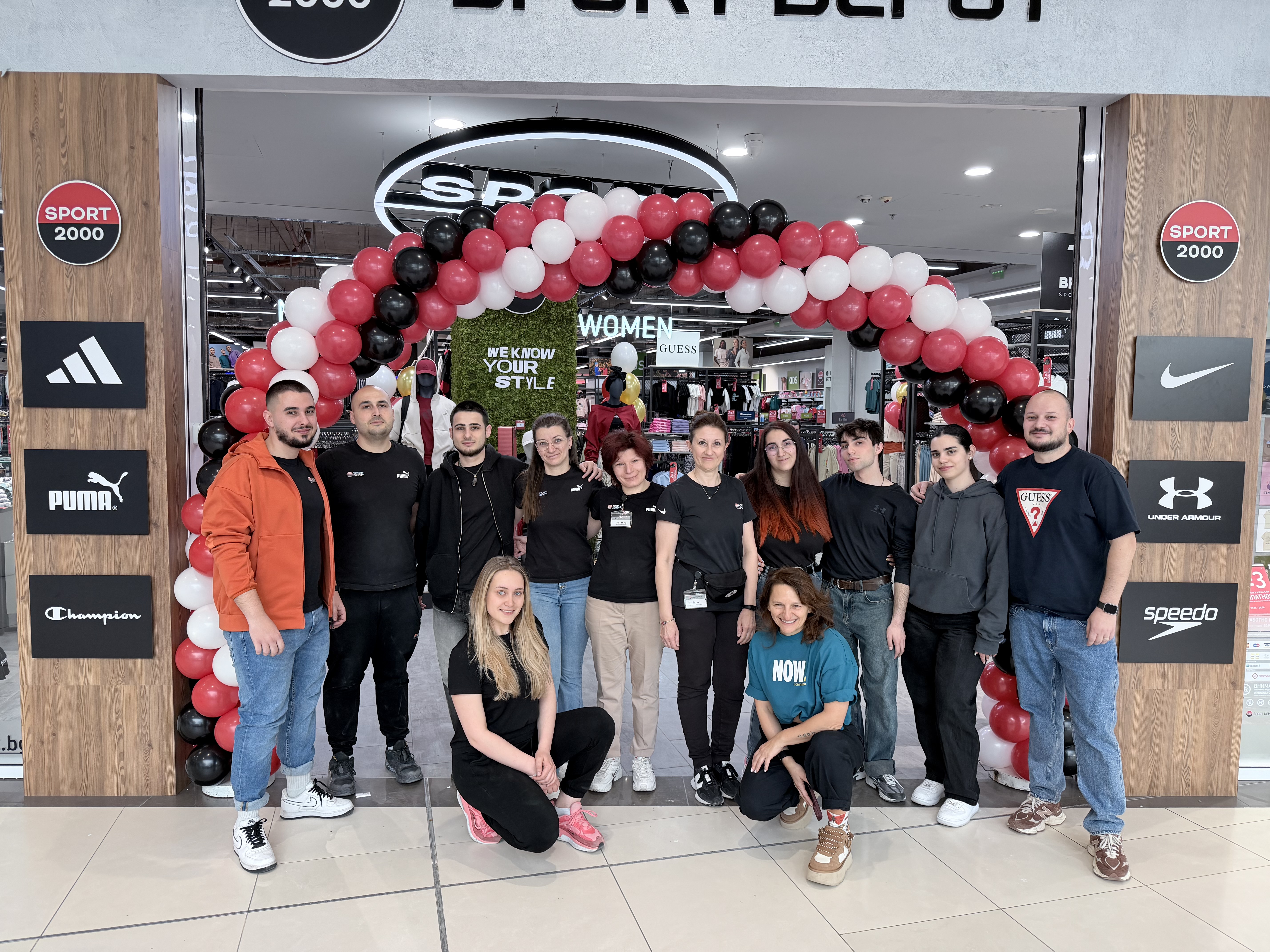 Обща снимка на екипа пред входа на обновения магазин, украсен с празнична арка от червени, черни и бели балони.