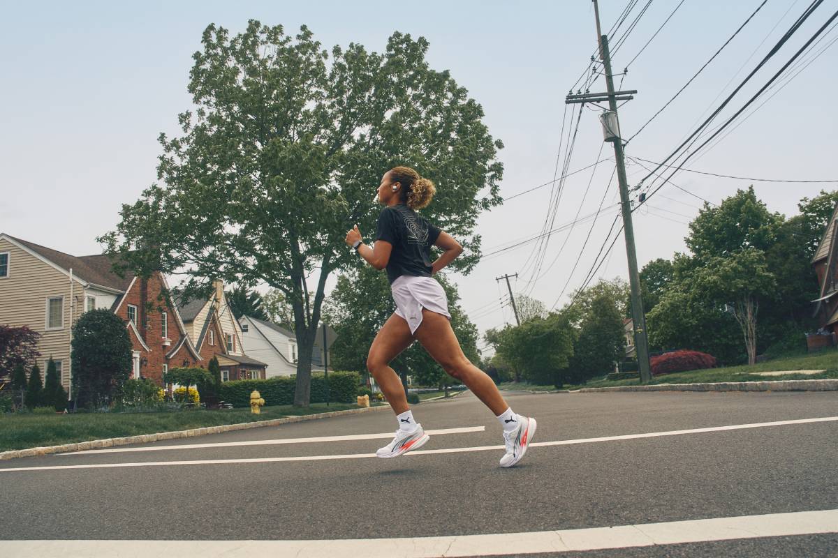 Жена бяга по улица в жилищен район, облечена в черна спортна тениска Puma, светли спортни къси панталони Puma и бели маратонки Puma Velocity Nitro 5.