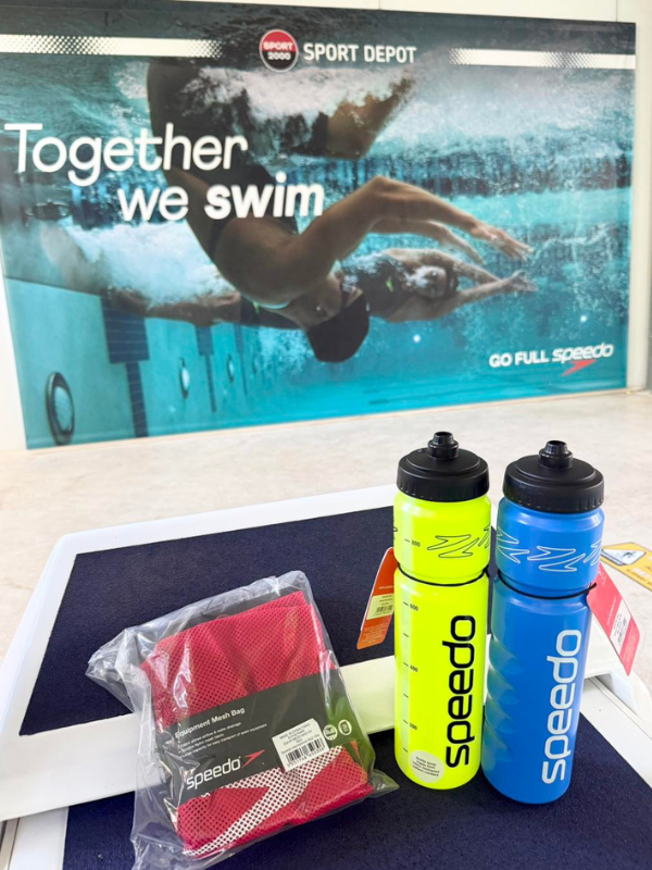 Плувни аксесоари Speedo &ndash; спортни бутилки за вода в неоново жълто и синьо, и мрежеста торба за екипировка, пред рекламен банер на SPORT DEPOT.