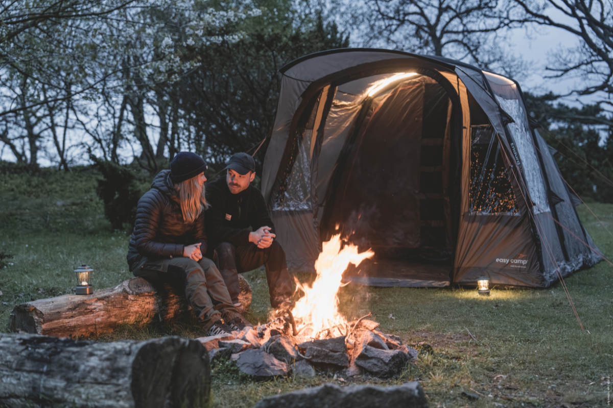 Мъж и жена седят около лагерен огън пред голяма къмпинг палатка Easy Camp вечер в гората.