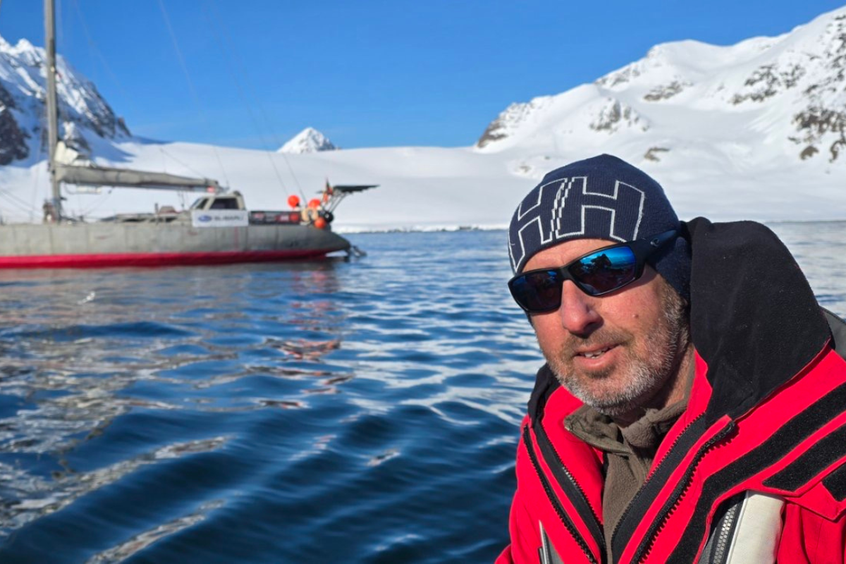 Член на Mirabo Adventures с висококачествено зимно яке и шапка Helly Hansen (HH), осигурени от SPORT DEPOT, на заден план с ветроходна лодка в ледените води на Антарктида.