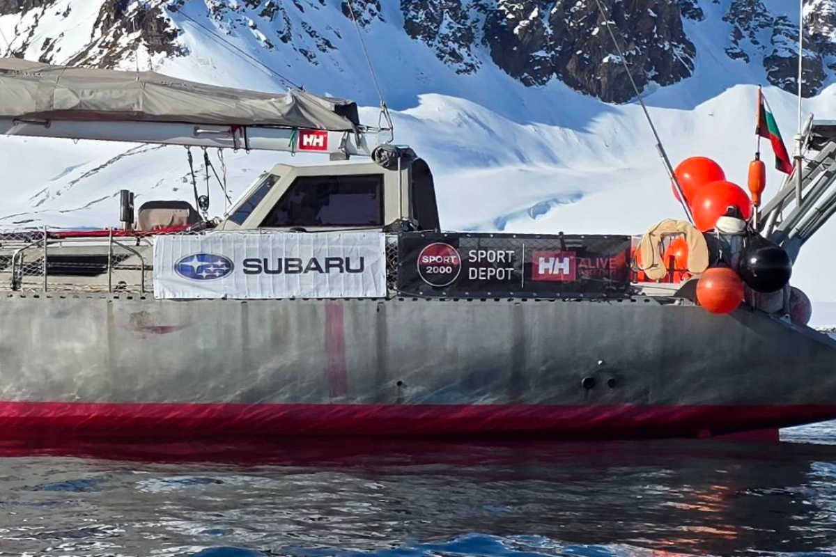 "Ветроходната лодка на Mirabo Adventures в Антарктида с рекламни пана на SPORT DEPOT, SPORT 2000, Helly Hansen (HH) и Subaru, закотвена пред заснежени планински склонове