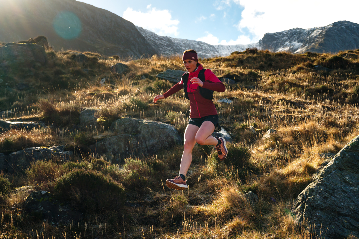 Жена тича по пресечен планински терен с екипировка за trail running в слънчево време