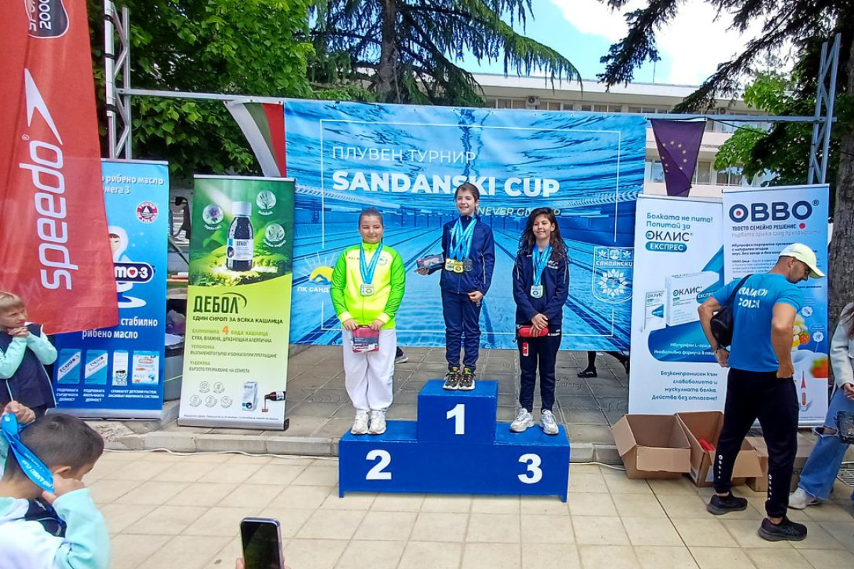 Три млади състезателки по плуване на почетната стълбичка по време на Sandanski Cup 2026. Момичетата са с медали на врата, застанали пред официалния банер на турнира. На преден план са позициите 1, 2 и 3, а около тях се виждат рекламни пана на спонсори, включително Speedo, предоставени от SPORT DEPOT.