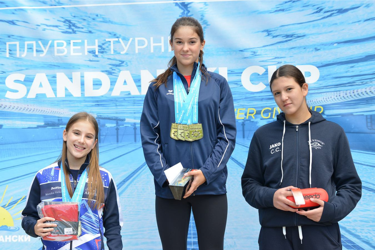 Три млади състезателки по плуване позират с наградите си след финала на Sandanski Cup 2026. Момичето в центъра носи четири златни медала, а състезателките държат аксесоари за плуване Speedo, предоставени от SPORT DEPOT.