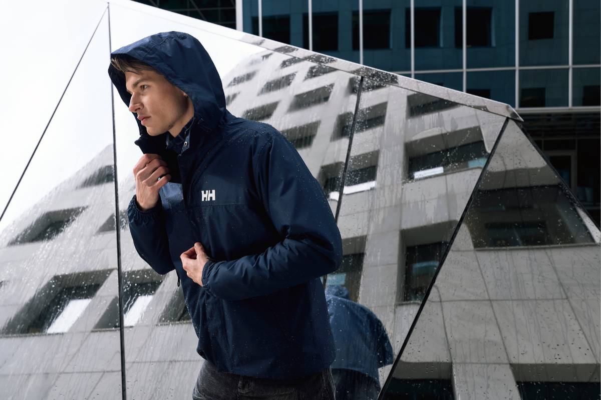 Мъж с тъмносиньо водоустойчиво яке Helly Hansen с качулка на градски фон.