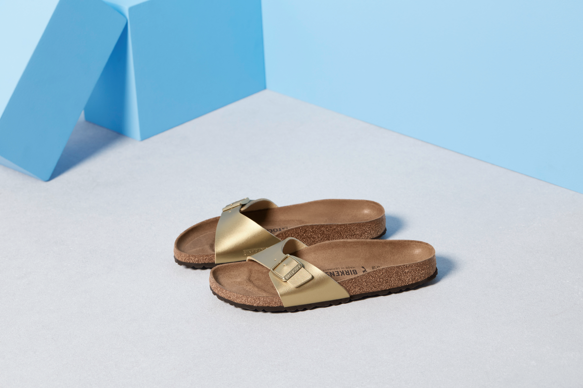 Златисти чехли Birkenstock Madrid с една каишка и коркова подметка, разположени върху сива повърхност на минималистичен син фон.