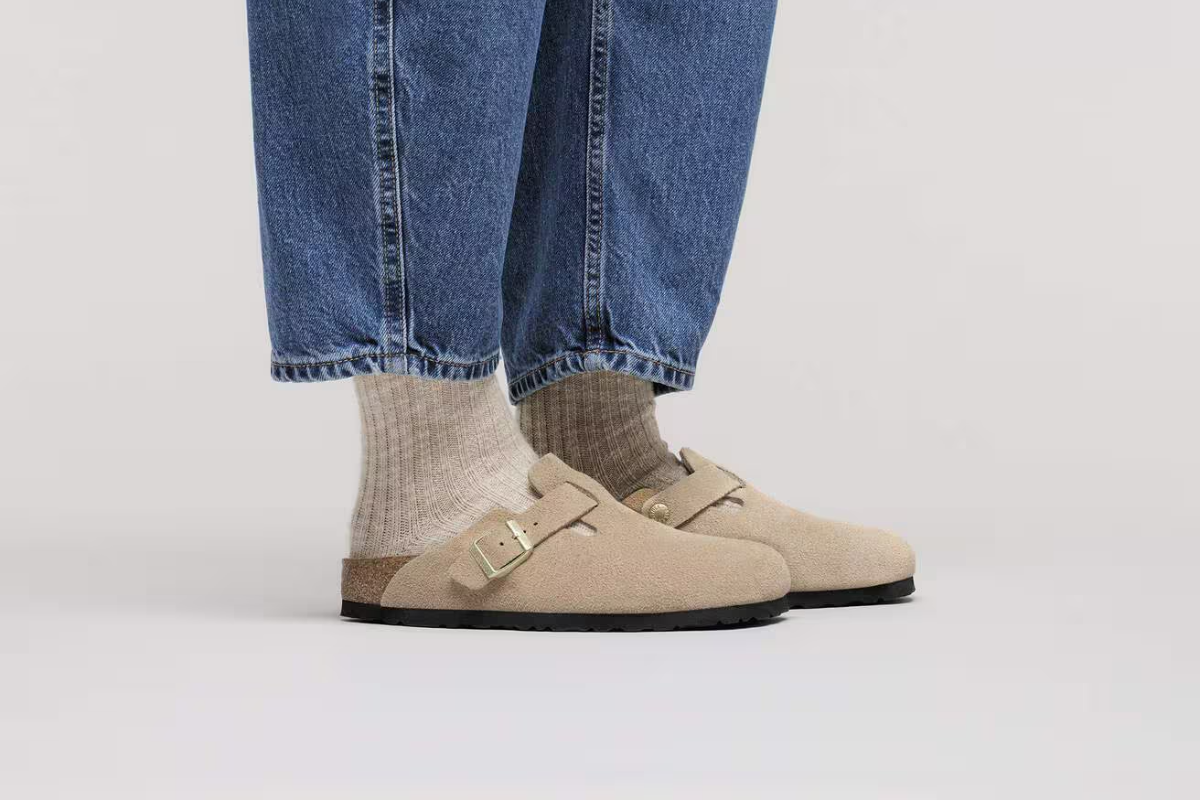 Човек, обут с велурени чехли Birkenstock модел Boston в бежов цвят, носени с плетени оребрени чорапи и сини дънки на сив фон.