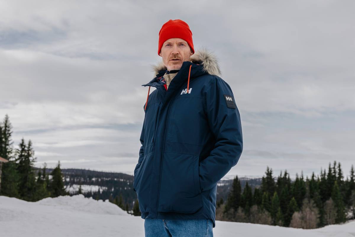 Мъж с червена шапка и тъмносиньо зимно яке Helly Hansen с пухкава качулка сред заснежен планински пейзаж
