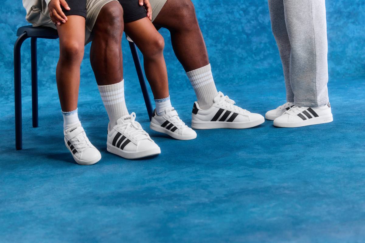 Краката на възрастен и деца, обути в бели кожени спортни обувки adidas с класическите три черни ленти, на син текстуриран фон