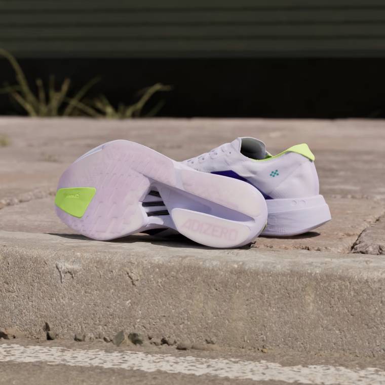 Изглед към маратонки Adidas Adizero Adios Pro 4 в бял цвят с три лилави ивици отстрани. Вижда се бялата подметка с пяна Lightstrike Pro и ярко жълт акцент на горната част на петата. Обувките са поставени на сив бетонен бордюр.