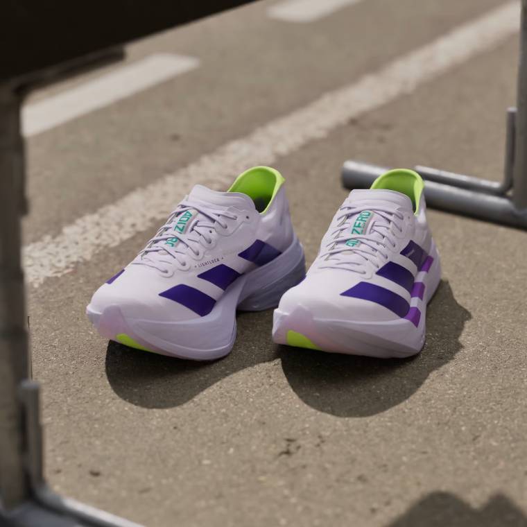 Изглед отпред на маратонки Adidas Adizero Adios Pro 4 върху асфалтова настилка. Моделът е с бяла горна част, три  лилави ивици, неоново жълти детайли и масивна бяла подметка.