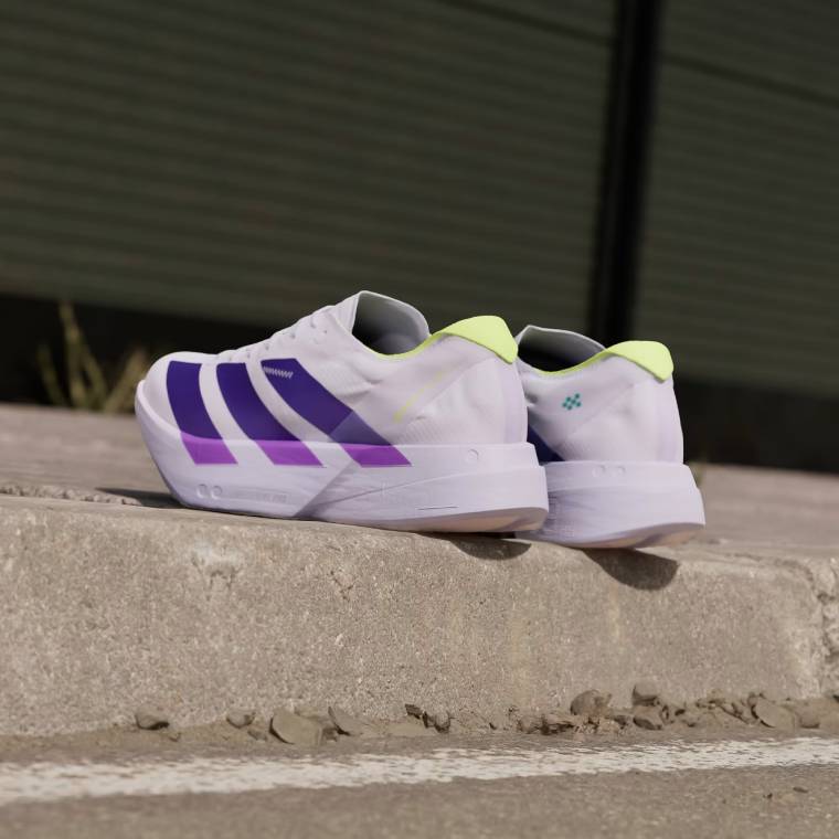 Чифт бели спортни обувки adidas Adizero Adios Pro 4 с три лилави ленти отстрани, флуоресцентно жълта пета и дебела бяла подметка, поставени на тротоар.
