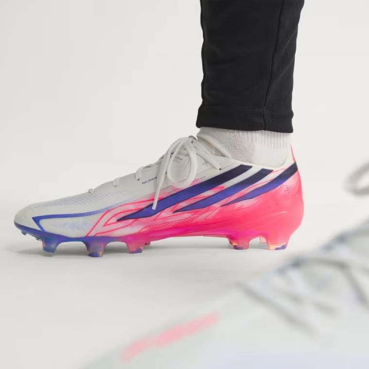 Близък план на бели футболни обувки adidas F50 Hyperfast Elite FG с розови графични детайли, сини акценти и цветна блестяща подметка с бутони за терен със суха естествена трева 