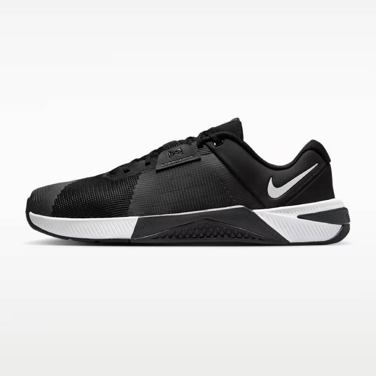 Профилен изглед на обувка за тренировки Nike Metcon 10. Обувката е с черно-бяла основа, дишаща мрежа в черен цвят, бяло лого Nike при петата и здрава подметка.